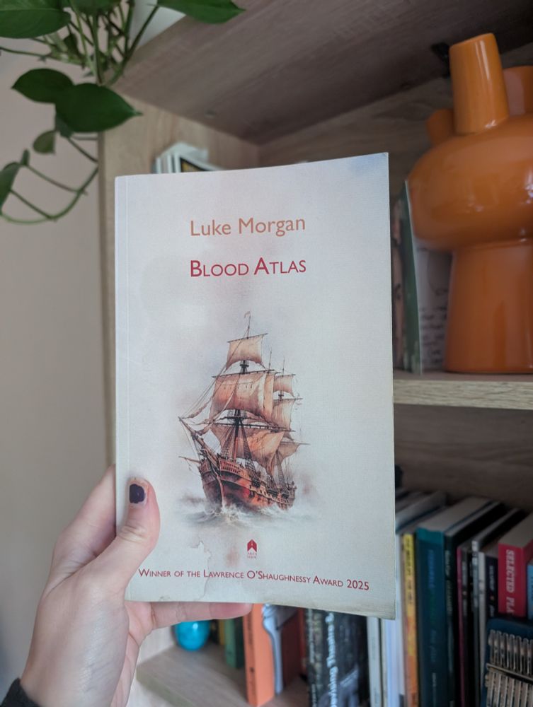 Cover of Blood Atlas is a large brown ship coming out of the ocean. The cover is cream.