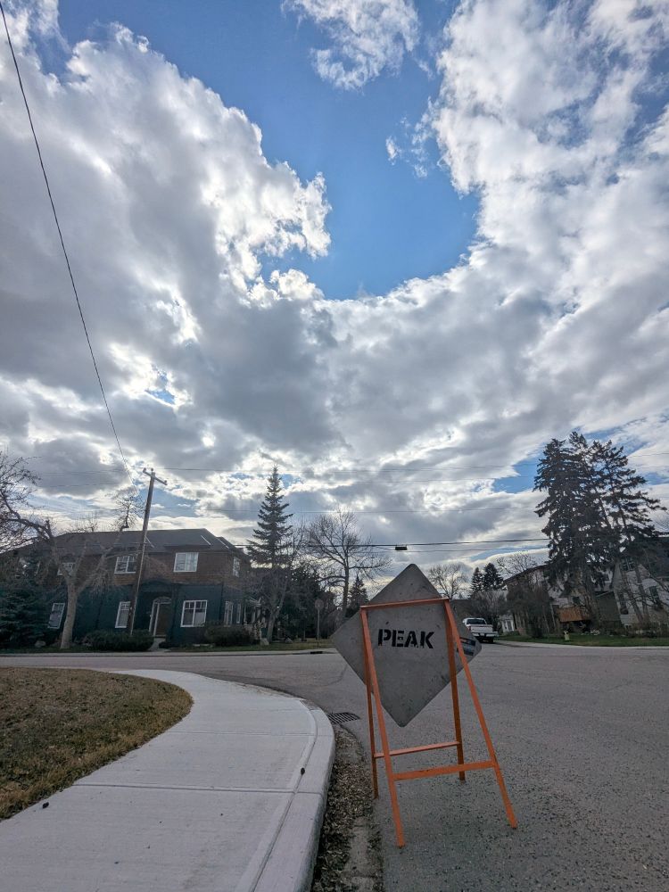 A residential street intersection featuring a geometric concrete sign reading "PEAK" mounted in an orange metal frame. The sign stands on a grass median strip between the sidewalk and asphalt road. Behind the sign, residential homes are visible, including a dark-colored two-story house on the left. Mature evergreen and deciduous trees frame the neighborhood scene. The sky displays dramatic cloud formations with a large circular opening revealing blue sky. Power lines cross horizontally through the upper portion of the image.