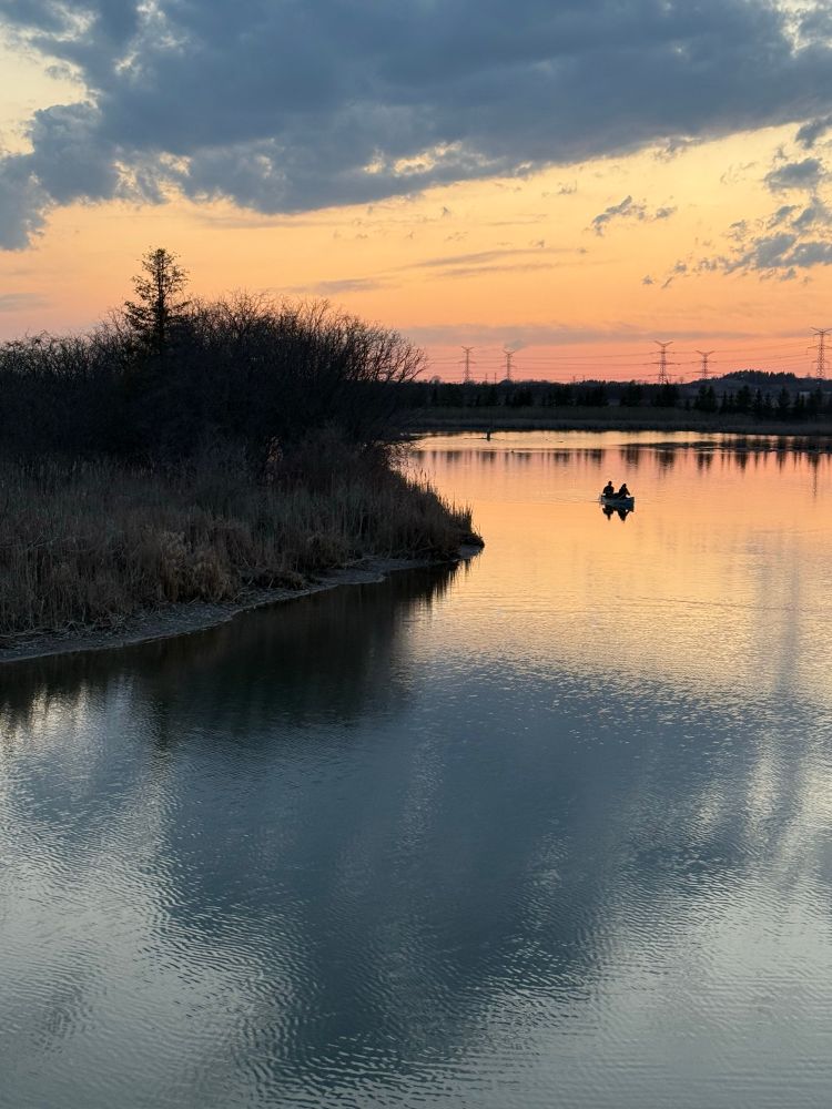 A picture of a body of water and the sky reflecting in it at sunset in shades of blue, gold, and peach. There is a spit of land covered in small trees, bushes, and grasses pushing out into the water on the left side of the image. Two fishermen in a boat row past the spit. 