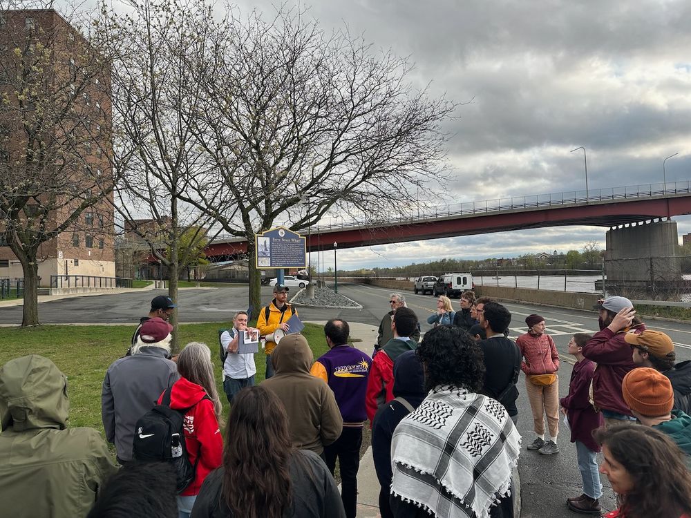 People gathered at the edge of the Hudson river, a man speaks into a megaphone.