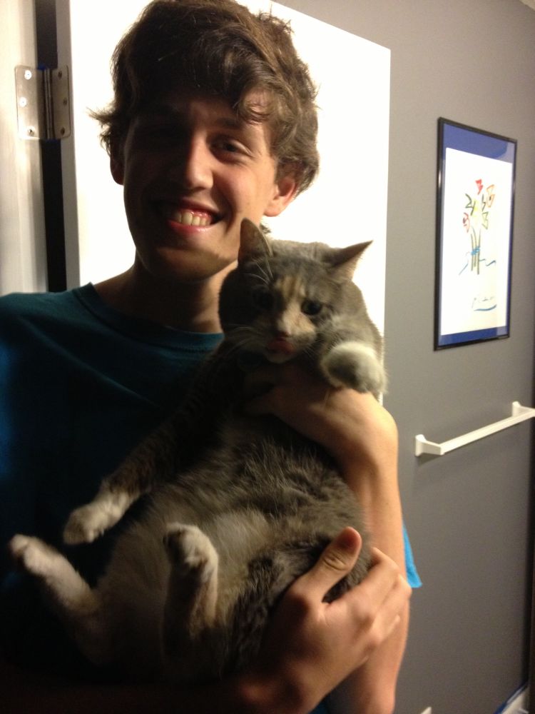 A smiling young curly brown haired man holds a cat who looks befuddled
