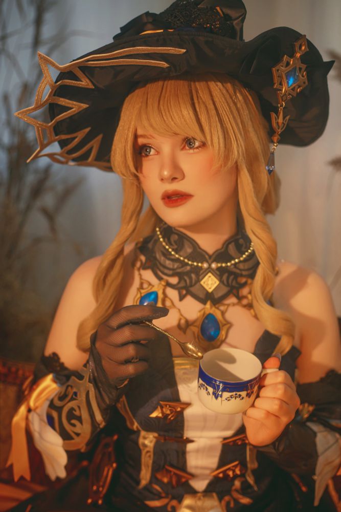 A cosplayer dressed as Navia from Genshin Impact poses in a warmly lit setting. She wears an elaborate black hat adorned with gold accents and star-like designs, complementing her golden blonde hair styled in loose curls. Her intricate outfit features dark and gold patterns, with blue gemstone details and delicate lacework around the neckline. She holds a blue and white porcelain teacup in one hand and a golden spoon in the other, her expression soft and thoughtful. The background is softly blurred, creating a serene and elegant atmosphere.