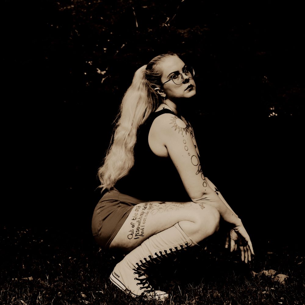 a blonde tattooed woman in shorts and knee high sneakers with long hair and glasses in sepia tone crouching and looking at the camera