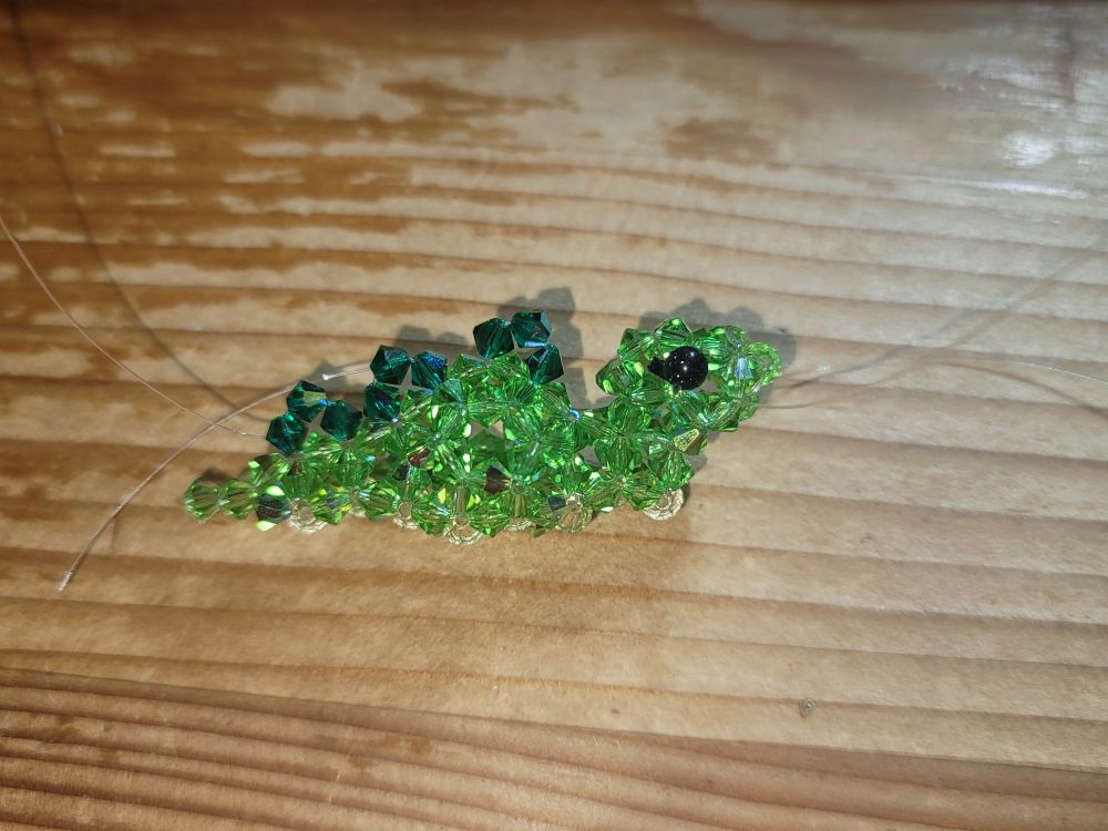 A half finished stegosaurus figurine made with crystal beads. Its body is light green, with dark green spikes on the back and a pale yellow belly. (Currently doesn't have legs because the dinosaur isn't finished yet)