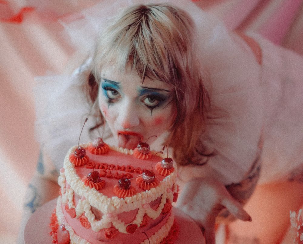 Daisy is wearing smeared dramatic clown makeup, bright blue eyeshadow and red lipstick. Daisy stares intensely at the camera while licking the top of a decorated pink cake. They are wearing a white frilly tulle garment, with blonde hair and a septum piercing. The cake is adorned with red cherries, ornate icing, and the word “Birthday slut” visible in icing letters. The background is soft pink and dreamlike, matching the surreal, theatrical aesthetic.