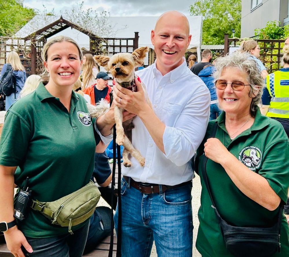 Picture of 3 people outside with a crowd of people walking around behind them. Max is in the middle of the photo, smiling widely, while holding Doodle the Yorkshire Terrier up to the camera. 