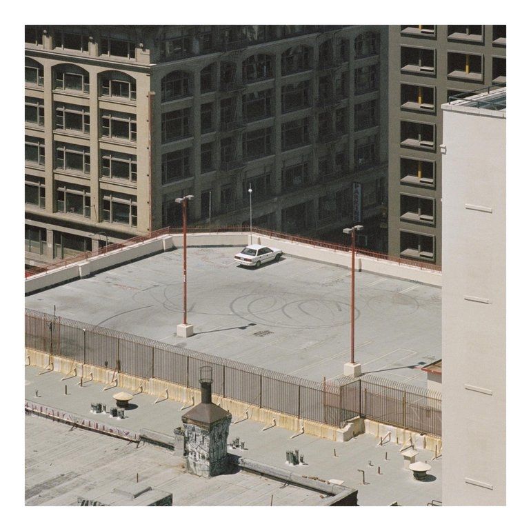 Cover art for Arctic Monkeys' album "The Car". It is a top-down, angled photograph of a car on a rooftop parking lot, framed with a white border. The photo is taken from far away by the band's drummer, Matt Helders, from his apartment. This distance really isolates the white car among the concrete of the surrounding buildings. Everything appears to be a few decades old, making the time period difficult to pin down. The colour palette is predominantly urban greys and browns, and there's something a little grainy about the photo. It has an isolating, liminal quality to it, like a random still from a film that was never released.
