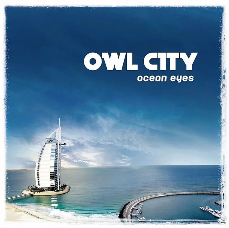 Cover art for Owl City's album "Ocean Eyes". It features a wide shot of the Burj Al Arab hotel in Dubai, overlooking the Persian Gulf. The stark white of the building blends somewhat into the clouds, which in turn blend into a deep blue sky. This is bordered in a speckled white, giving the impression of being a painting on a canvas. The album and artists name are placed near the top right in white lettering. The photograph looks hyper real; edited to sell a feeling of adventure and open ocean, not unlike what a marketing still would do.