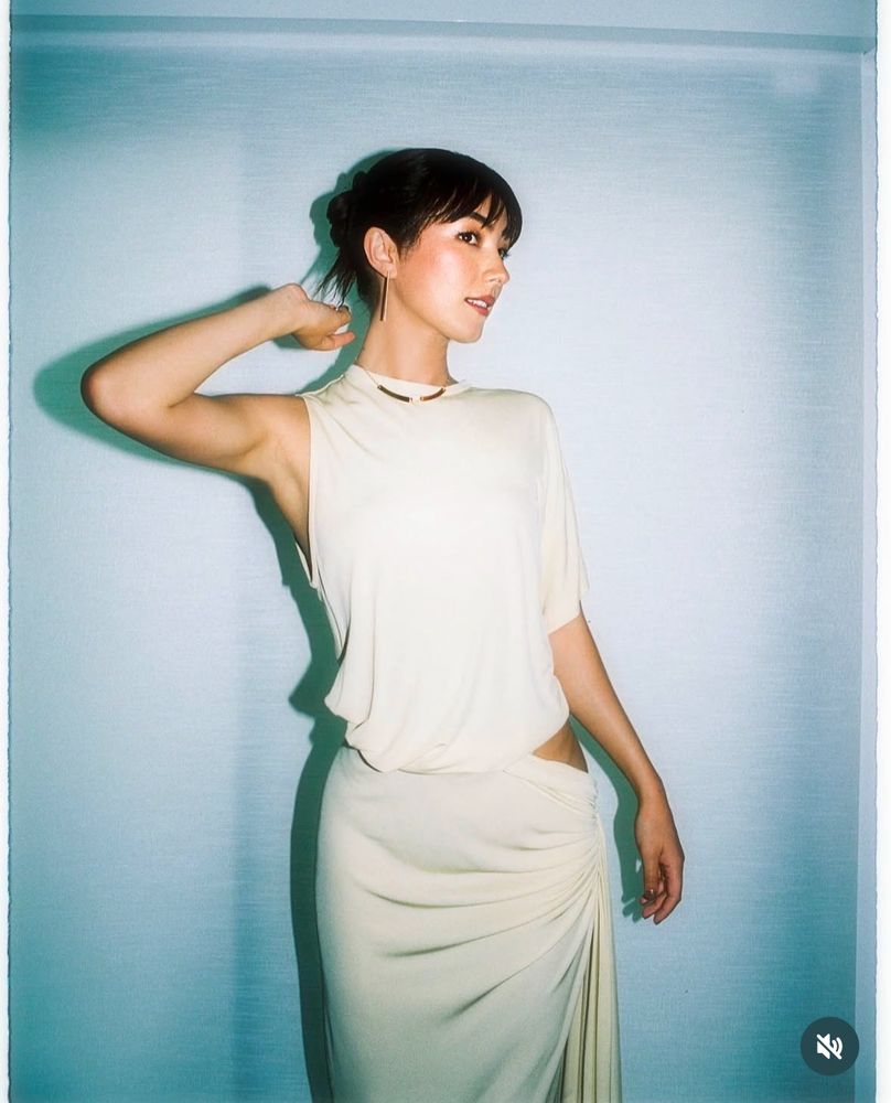 A photo of Natasha Liu Bordizzo wearing a lovely cream dress as she poses for a photo. It is sleeves on the left while the right has a sleeve that goes to her elbow. Her hip is also exposed a little on the right side of the dress.