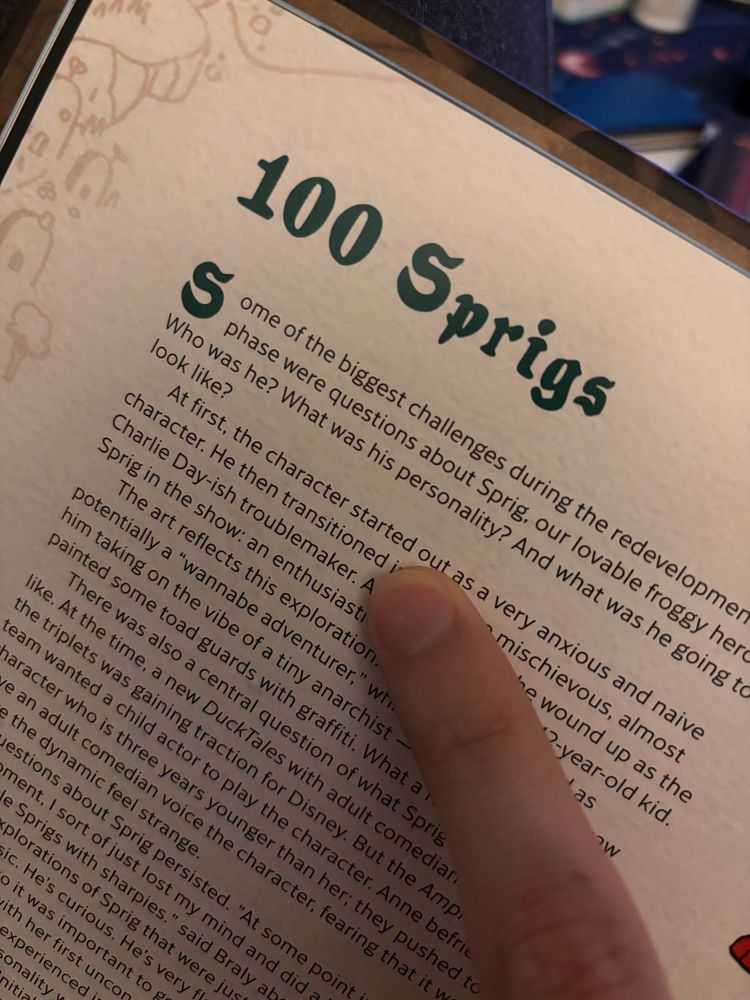 A page from the Amphibia art book. The title is “100 Sprigs” and a finger is pointing to the text: “He then transitioned…”