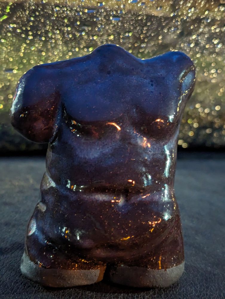 Photo of a sculpture taken at night in front of a rain-spattered windshield. The sculpture is of a plus size trans masc torso in black clay with an opal glaze over it that leaves most of the body dark and glossy, but settles in the recesses as a pearly color. The glaze highlights where top surgery scars would be. 