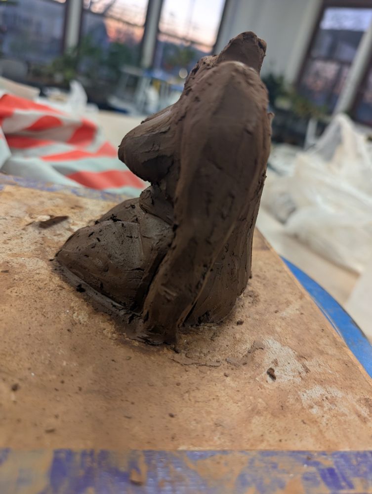 A side view of a headless dark brown clay figure of a femme body leaning forward. It sits on a clay-streaked board in front of open windows. The sun is setting in the background. 