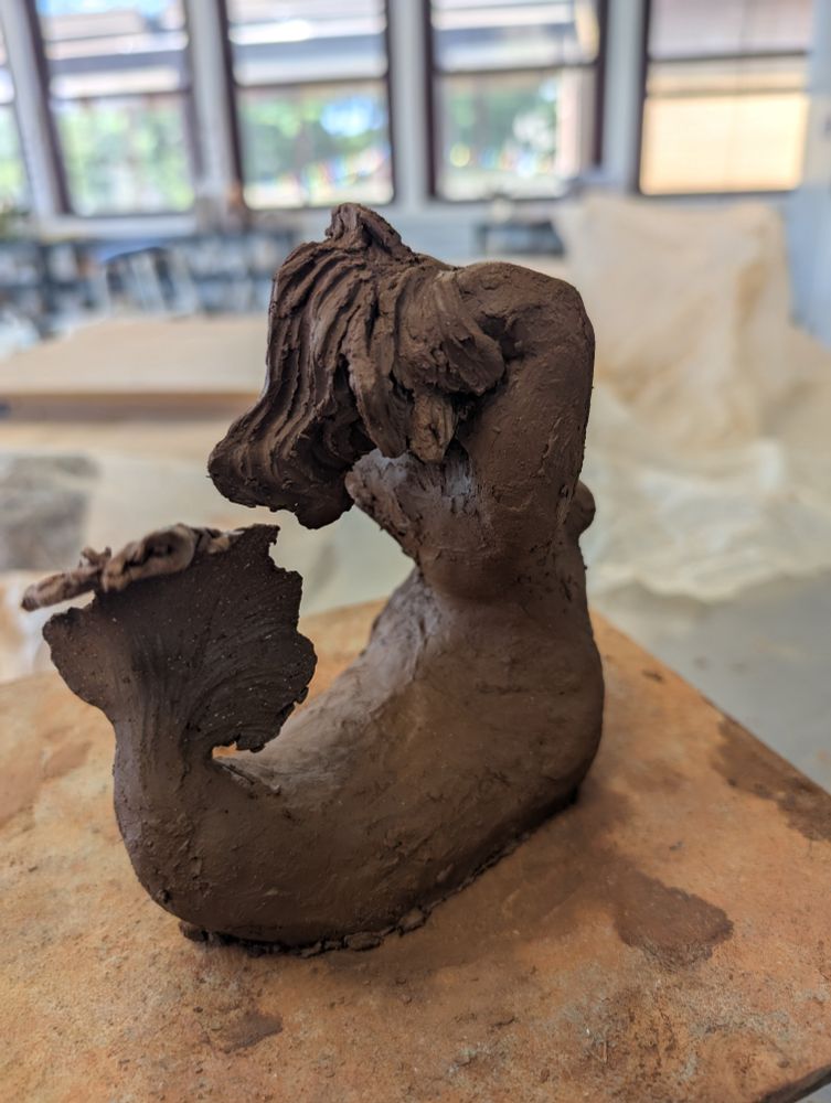 A sculpture of a mermaid from the side, leaning back with her hands in her hair and her face turned upward. Behind her is a crumpled piece of plastic and the pottery studio she was made in. 