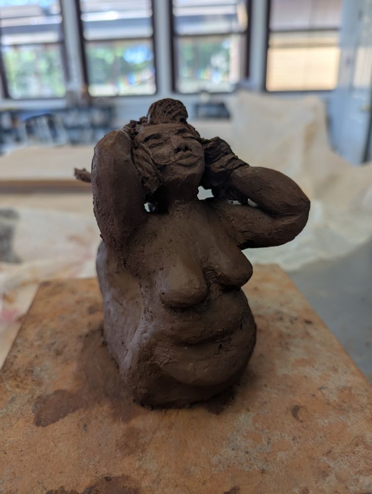A sculpture of a mermaid from the front, leaning back with her hands in her hair and her face turned upward. Behind her is a crumpled piece of plastic and the pottery studio she was made in. 