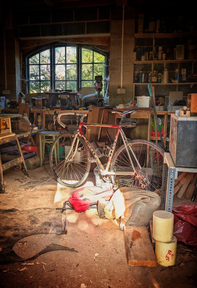 A racing bike sits in a cluttered and dirty workshop, illuminated by a golden ray of sunshine.  A sunny day is visible through an arched window in the background