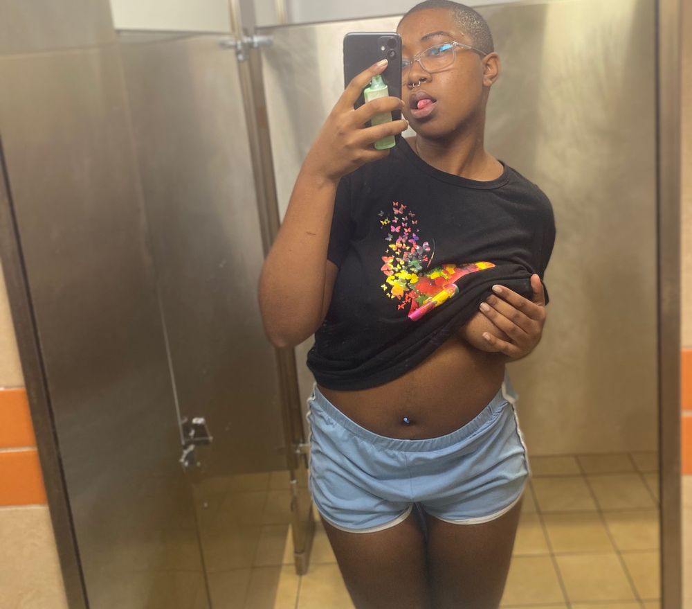 Naomi taking a selfie in the mirror blue shorts white border bottom and black butterfly shirt lifted on the right hand covering exposed part, and belly button and belly ring showing