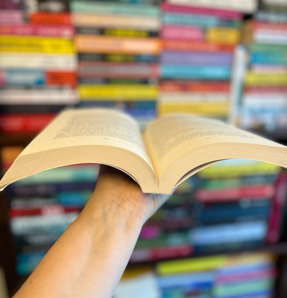 A book open, held on one hand in front of a blurred bookcase full of books 