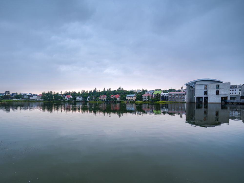 A wide-angle view of Tjörnin, the central city pond in Reykjavik, Iceland, on a calm, overcast day. The water is still, mirroring the sky and the row of buildings that line the far shore. Most of the buildings are modest, painted in soft whites, reds, greens, and grays. On the far right stands the curved, concrete form of Reykjavik City Hall, partially elevated above the water. Above the skyline, the sky is pale and cloud-covered, casting a gentle, muted light over the scene. The atmosphere is peaceful, almost meditative.