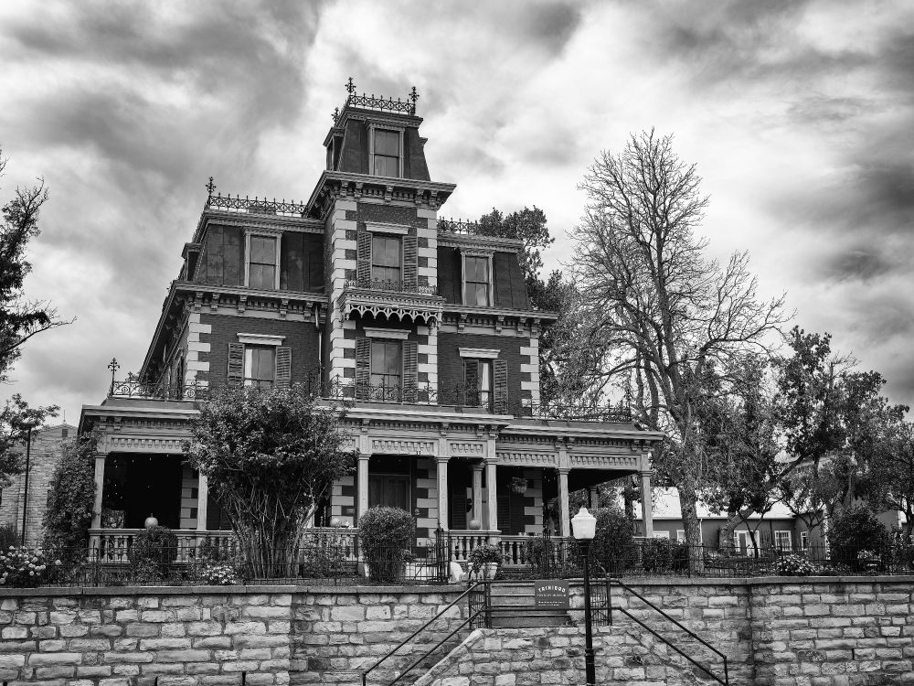 A grand three-story Victorian home dominates the frame in striking black and white. The building’s red-brick façade (rendered in gray tones) features decorative cornices, tall shuttered windows, and a central tower capped with intricate ironwork. A stone retaining wall and staircase lead upward toward the porch, where carved columns and spindled railings accentuate the structure’s symmetry. The trees around it are uneven—some leafy, some bare—adding a spectral quality to the scene. Heavy clouds swirl above, echoing the mansion’s ornate drama.
The photograph evokes both elegance and unease—a preserved relic standing proud against time, haunted not by ghosts but by its own persistence.