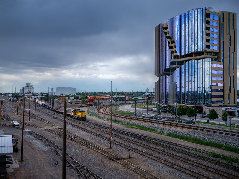 A dramatic cityscape showing the convergence of a train yard and a striking modern high-rise. On the left, multiple sets of freight tracks stretch into the distance under a dark, brooding sky. A yellow Union Pacific locomotive sits idle in the foreground, facing away from the viewer. Farther back, colorful railcars fill the switching yard. On the right, a tall mirrored building reflects a distorted sky—its glass façade warped with deep sculptural curves, contrasting sharply with the industrial sprawl. Storm clouds press down from above, softening the light and lending the scene a tense stillness.