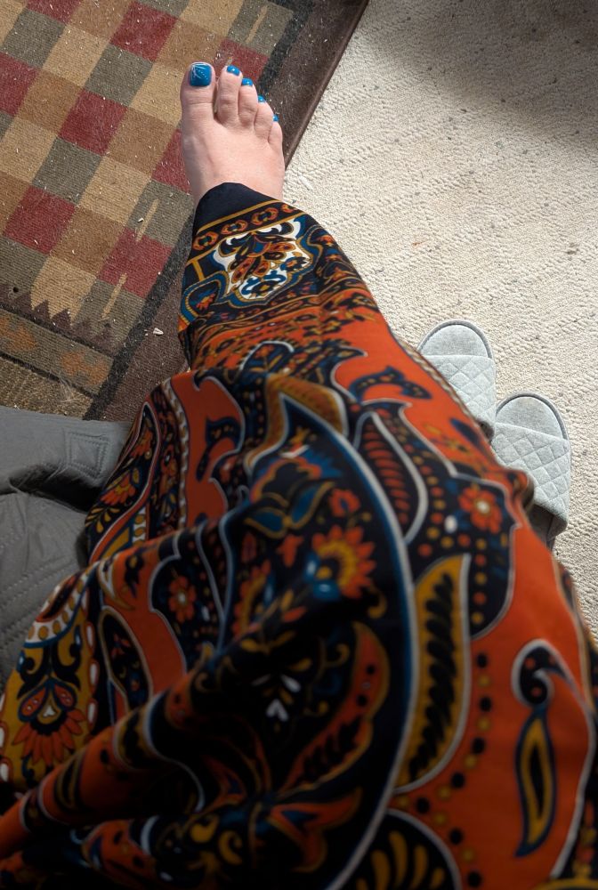 Looking down at a right leg crossed over the left, draped beneath a paisley patterned caftan in shades of dark blue and burnt orange. The toenails of the right foot: sky blue. The carpets beneath the foot: Filthy, gross, omg I have to vacuum tomorrow.