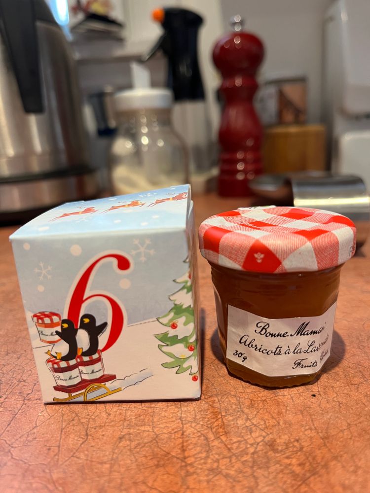 A small box is on the left, ,with a red number six on it, and two penguins on a sled. The sled has three jars of jam on it as well, tiny versions of Bonne Maman jars. On the right is a small jar of actual jam, with a checkered red and white lid. The label has a handwriting font and says: Bonne Maman, Abricots à la Lavande - 30g - Fruits Choisis (Apricot and Lavender) 