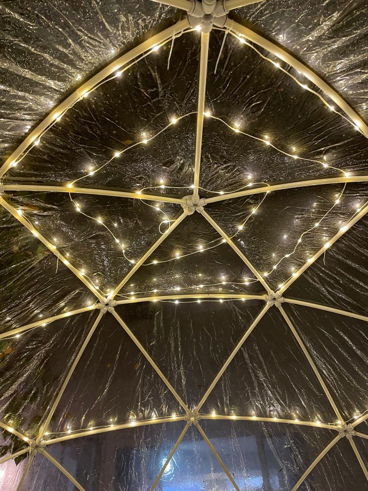 Looking up from the inside of a transparent bubble dome with white mini-lights in a spiral pattern strung along the white PVC pipe frame. 