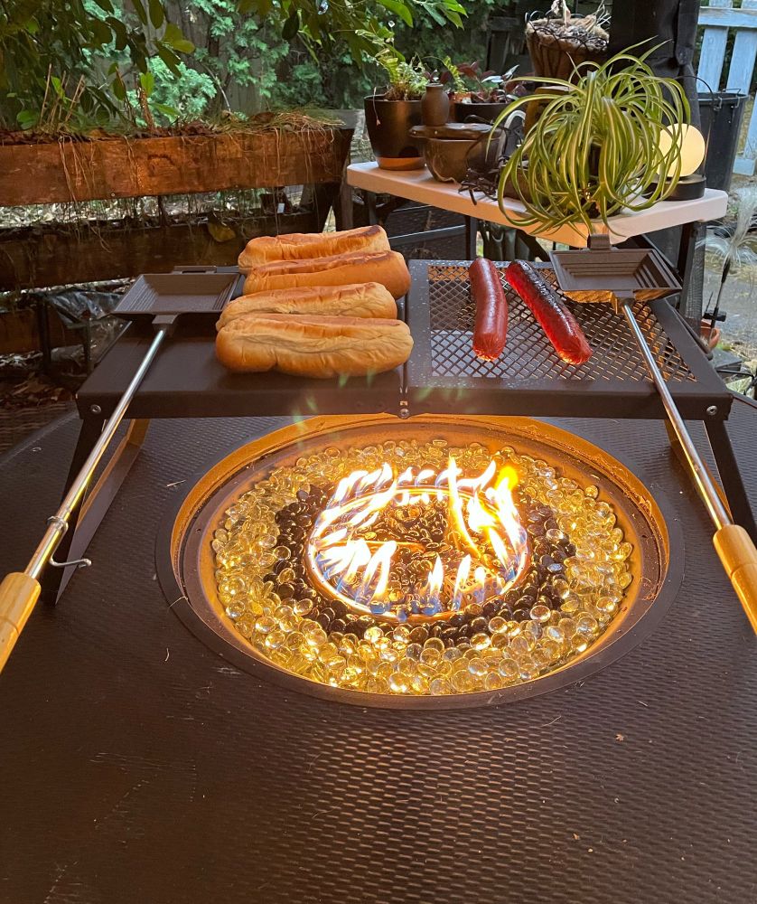 Hot dogs and hot dog buns sitting on top of a cooking grate on a propane fire table. 