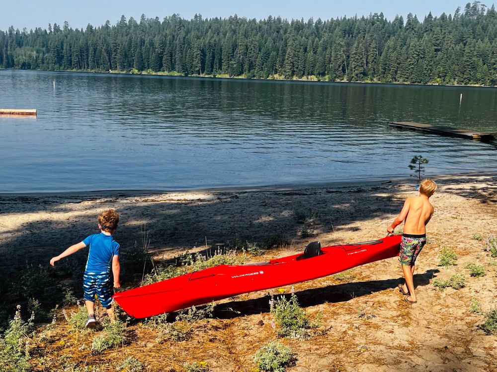 Two young boys carrying a big red kayak down to a lake. 