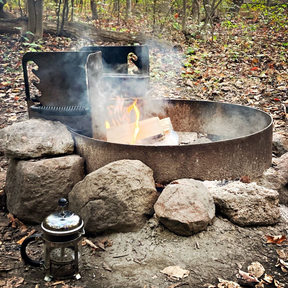 An old campfire metal pit surrounded by stones burns with a strong fire. On the camp grill is a pot, and a French press sits open nearby.