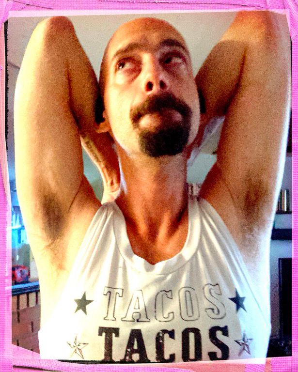 A person with a shaved head and goatee is wearing a sleeveless shirt and exposing their pits, hands behind their head. They have a mischievous expression and are looking off to the upper left of the frame.