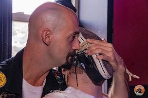 A masculine person with a shaved head leers behind a man, covering his face with a converse shoe.