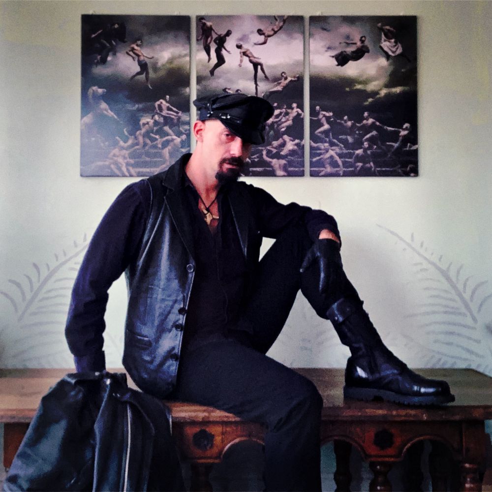 A masculine person with a goatee dressed in all black sits on an altar in front of a triptych of Renaissance male poses. They are wearing tight pants, formal leather boots, leather gloves, a leather police cap, and a leather vest. On the altar next to them is a leather biker jacket.