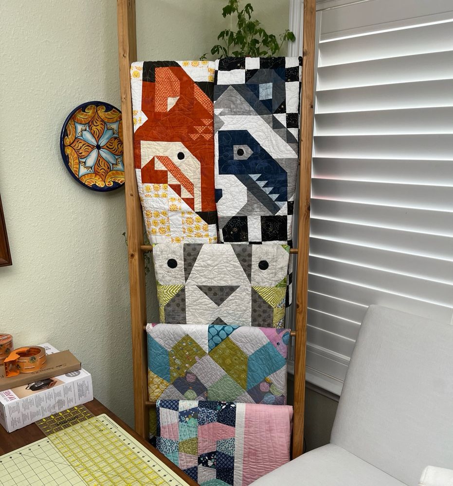 Quilts displayed on a ladder: bottom row pink, white, aqua, and navy tumbler, second row sage, gray, aqua, yellow, and fuchsia, third row medium gray, off white, light gray, two black circles (a very simple bear face), top row is two quilts on the left a fox face, on the right a raccoon face. The faces line up. In the foreground is a white chair and a dark wood table with a cutting mat and ruler. In the background is an off white wall, white shutters, a decorative plate in blues and gold, and a plant 