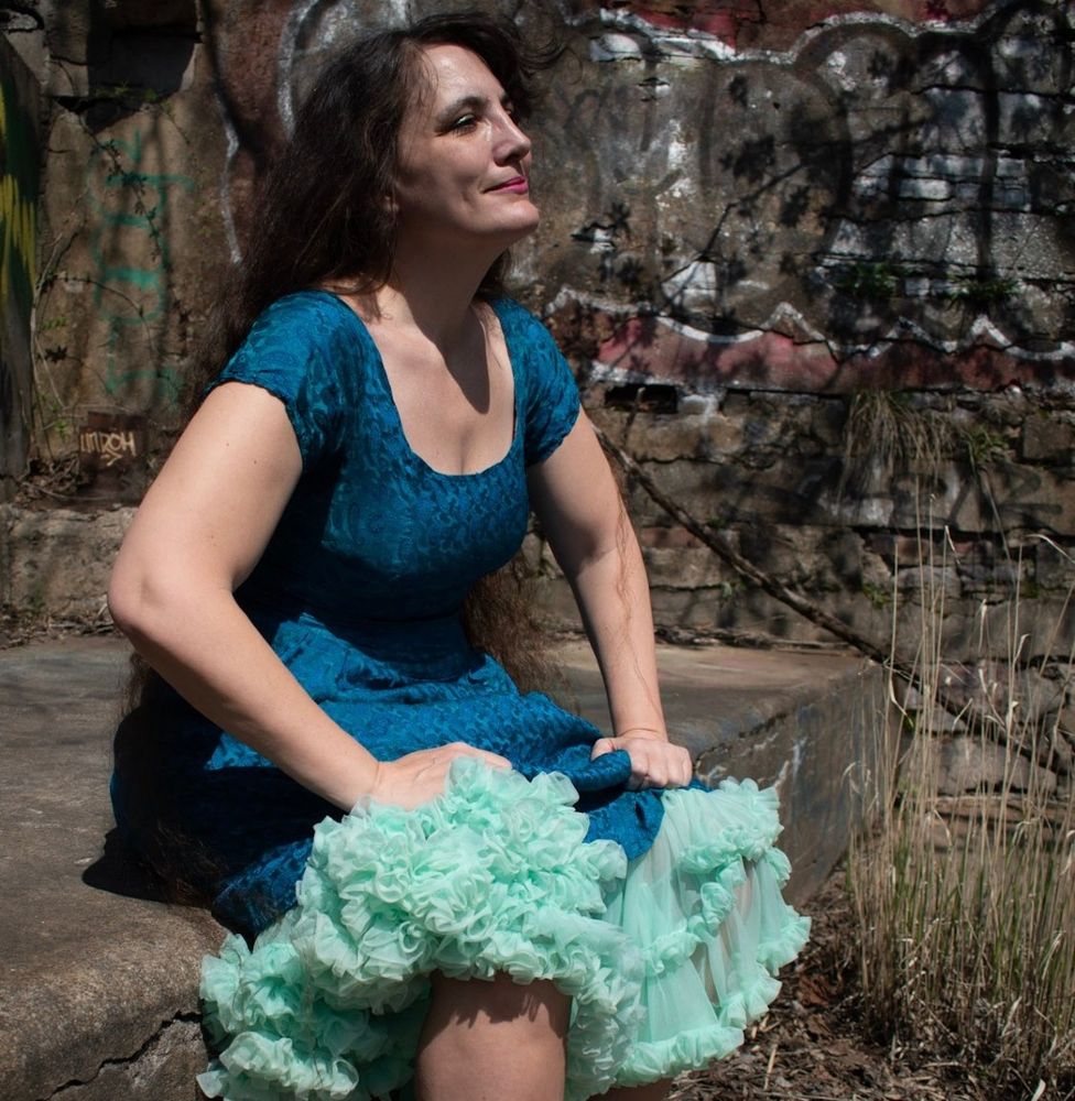 Woman with pleased smile sits with legs wide and puffy skirt ruched up. 