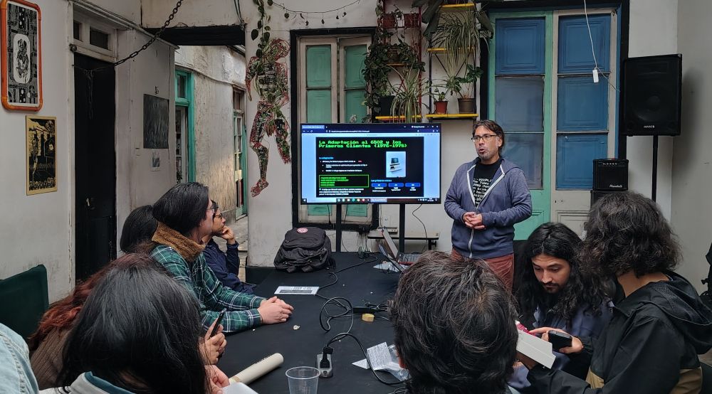 fotografía amplia del interior, pantalla al centro, mesa con varios aparatos electrónicos, expositor de pie y varios oyentes sentados