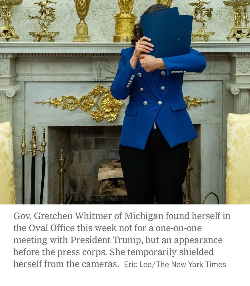 Photo of Michigan governor Gretchen Whitmer standing in front of the fireplace in the Oval Office in a blue blazer hiding her face from the cameras by holding up blue file folders. 