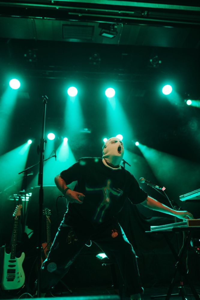 POULISH KID, wearing a corn-themed mask and a colorful outfit, moves energetically across the stage while playing the synthesizers.