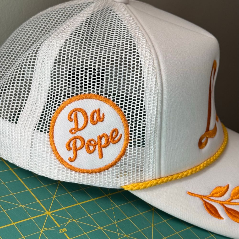 Side view of hat. White patch with gold border sewn onto mesh side panel. Circular. Da Pope is embroidered in same script as Leo. 