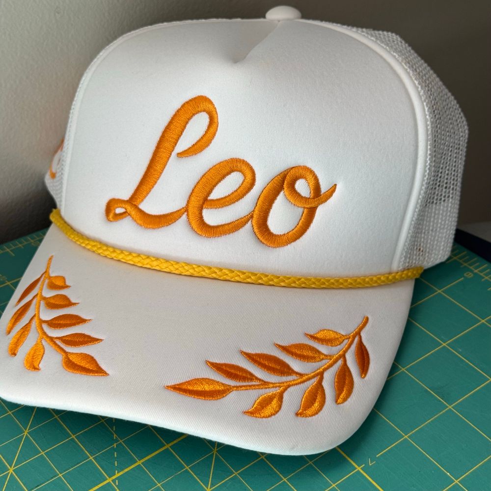White foam trucker hat. Bill embroidered with gold laurel leaves similar to the “scrambled egg” design on some military caps. Yellow braid across the top of brim. The name Leo, written in script, is embroidered in gold on the front. 