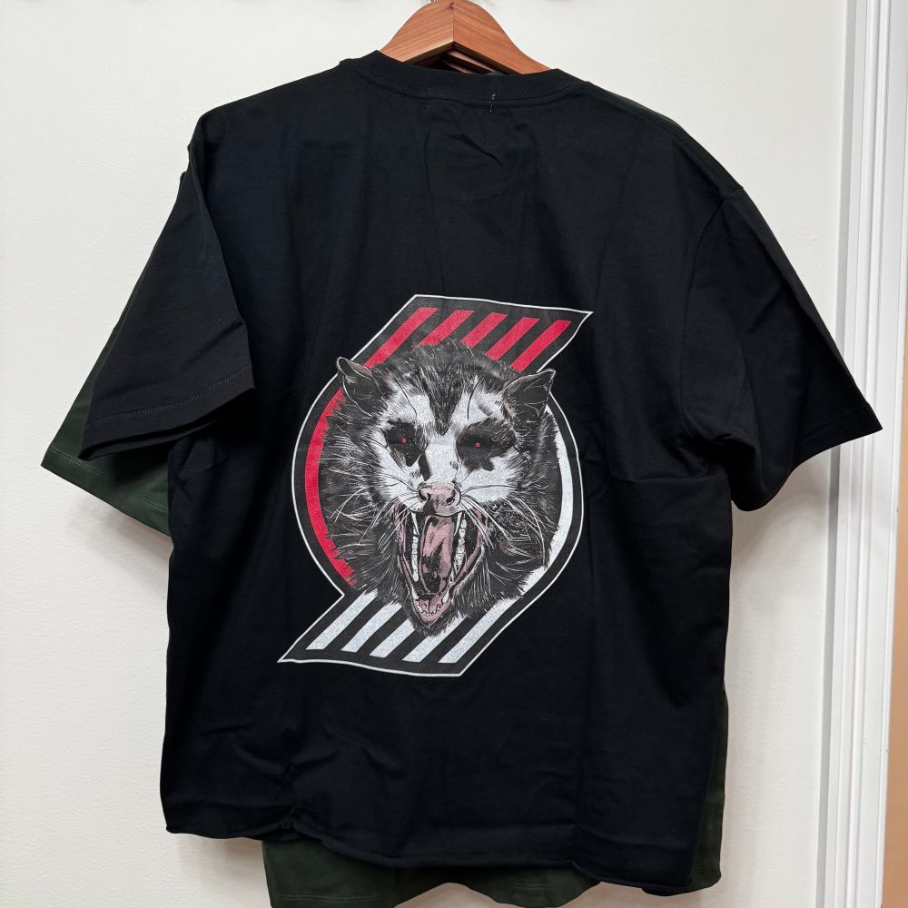 Back of acabcity tee. An illustration of a possum with fangs bared and glowing red pupils looking out at the viewer. Behind the possum is a design of a red and white pinwheel