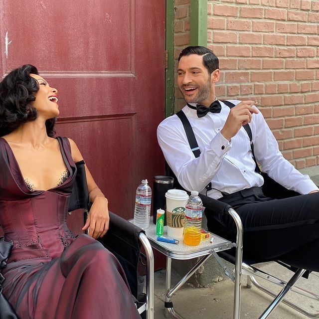 A behind the scenes shot from the TV Show Lucifer of Leslie-Anne Brandt, wearing a burgandy, 1920s evening gown, laughing as she sits next to Tom Ellis, wearing black trousers, a white shirt and black bow tie