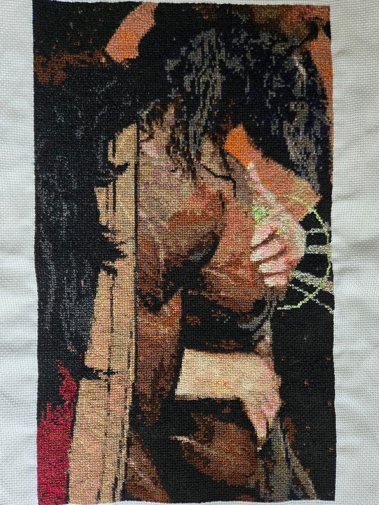 A photo of a cross stitch project on white aida fabric. The pattern is based on Blackwall's romanced tarot card design from Dragon Age Inquisition. A shirtless man with long dark hair and a scraggly beard is wearing a red and gold cloak with black fur. There is a moon behind him. A woman's hands are coming around from behind - one is on his chest over his heart and glows green.
