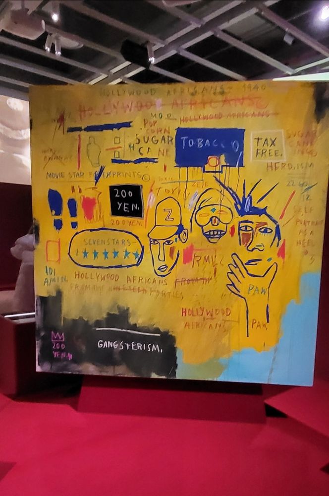 Abstract grafitti art work that is mostly yellow with drawing of characters in baseball caps, sunglasses, text written all over Museum text: The entertainment industries of music, Hollywood, and theater were important pathways for Black artists to make a living. These opportunities, however, often forced them to confront, subvert, and redress histories of stereotypical representations and exclusion, a reality grappled with by Jean-Michel Basquiat through the crossing out of words...