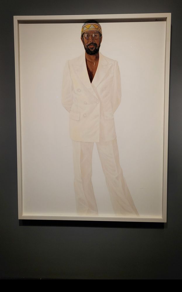 Painting of a grown Black man with brown skin and a well-groomed moustache beard, who is wearing a kufi (Afrocentric skull cap), stylish glasses, and an impecably tailored white double-breasted white suit. The trousers draped beautifully. He is wearing a chain with a (flexed leg ?) pendant and no shirt. It's against a white background. Elegant and attractive Black people in white-on-white is a Hendricks trademark.
Museum text:
Barkley Hendricks's self-portrait Slick is an iconic image of a stylish Black man in the 1970s who expresses himself and cultural confidence with aplomb. Looking straight at the viewer in an impeccably tailored, white double- breasted suit, Hendricks wears a kufi, a woven skullcap of African design often donned by Muslim religious figures and elders within the African diaspora, similar to the example shown here. According to the artist, the kufi was included to "enhance the composition," the colorful fabric amplifying the contrast. These elements plus the lightly tinted glasses, gold necklace, small gold hoop earing. and toothpick communicate Hendricks's dedication to beauty, cool, and heritage.
#BlackSky #ArtSky