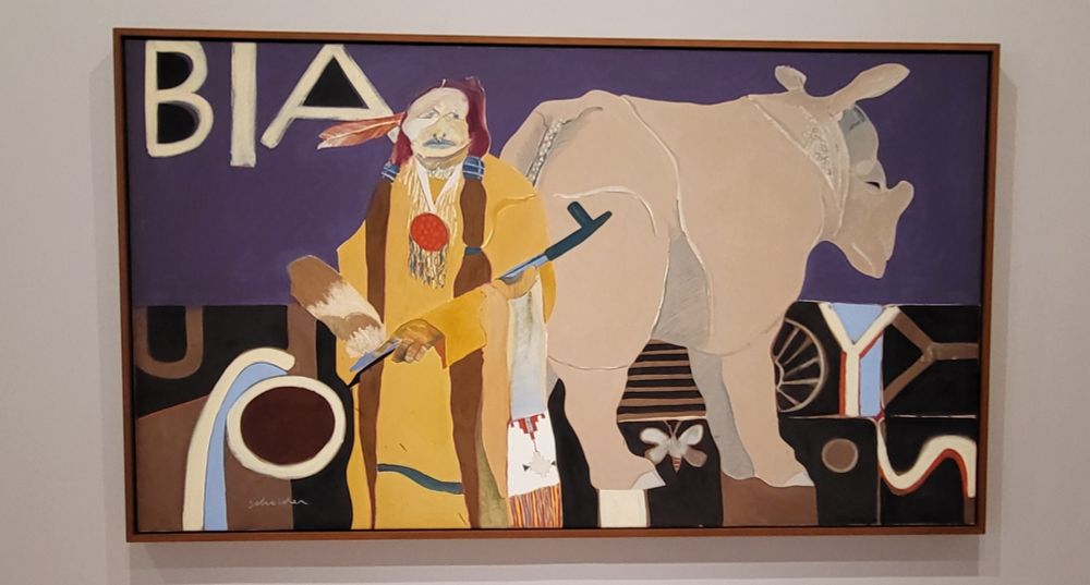 short description: painting of a Native American in full native dress with a peace pipe standing in front of a rhinoceros  standing next to BIA (Bureau of Native Affairs)
by Fritz Scholder, American and Luiseño Band of La Jolla Indians, 1937-2005.

Museum text: "My painting called Indian and Rhinoceros has an Indian in full dress with a peace pipe posing with an ungainly rhinoceros in front of the Bureau of Indian Affairs [BIA]. My point,I believe, is obvious." An enrolled member of the Luiseño tribe, Fritz Scholder's statement underscores the cruel absurdity of the US government's relationship with Native Americans. The BIA, an agency within the Department of the Interior, was responsible for enforcing decades of federal policies designed to terminate tribal authority and relocate and assimilate Indigenous people. Galvanized by the escalation of he Vietnam War, in which Native American men disproportionately served, and the 1968 founding of the American Indian Movement (AIM), an activist group focused on Native American civil rights, Scholder felt compelled to address the violence of American history and the mistreatment of lndigenous people for the first time in his work.