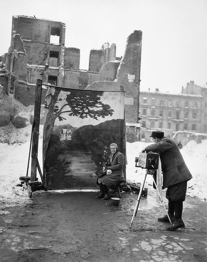 Black and white photo of a photographer taking a picture of a seated smiling woman in front of a fake backdrop depicting a scenic road leading into town while the same road and town is just behind them, completely bombed.