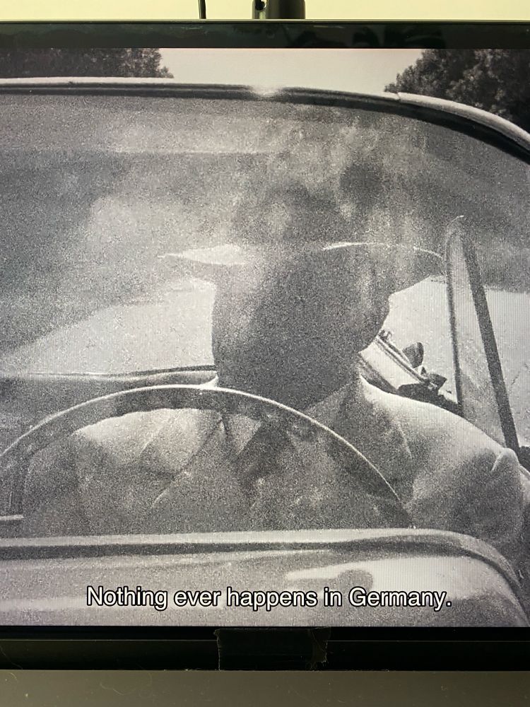 Film still from Rainer Werner Fassbinder, The American Soldier, 1970. Karl Scheydt, wearing a suit and a hat is behind the wheel of a convertible. His face isn't recognizable through the windshield reflecting the sky. Below, the subtitles read: "Nothing even happens in Germany."