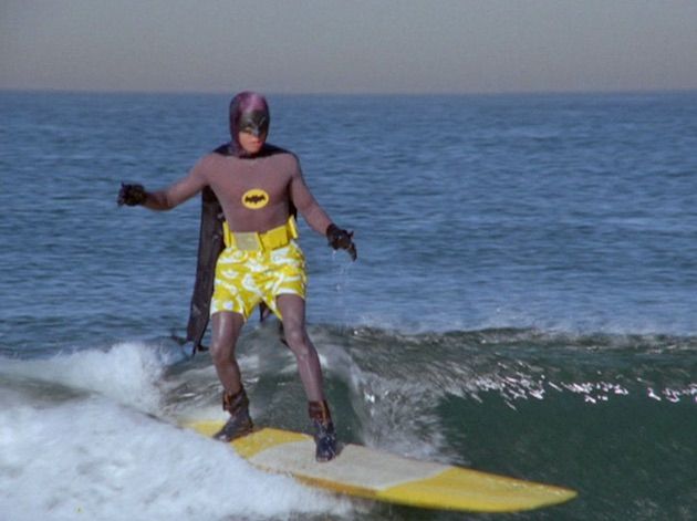 Batman in his suit with yellow shorts over the top, riding a wave on a surfboard