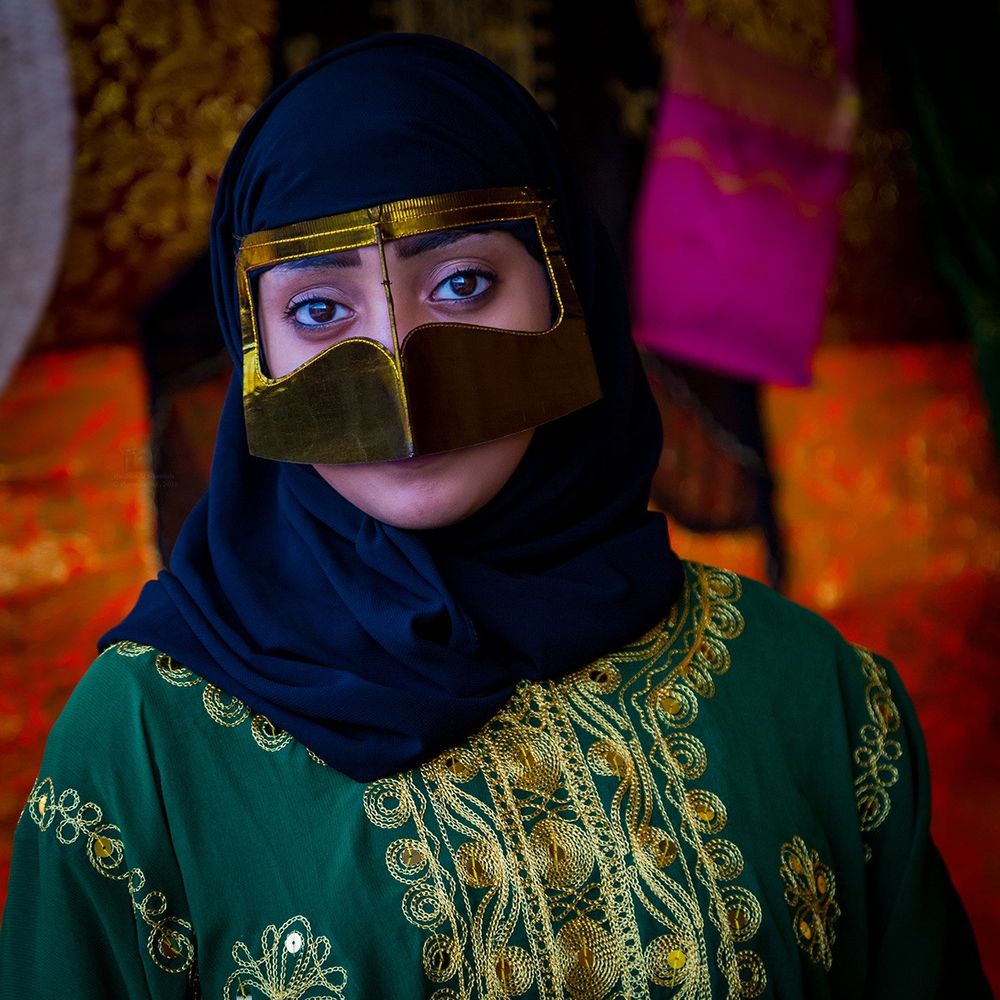 Woman in a green dress and black headscarf with a golden battoulah, a metallic knight's-visor-looking mask that frames the eyes and covers the mouth.